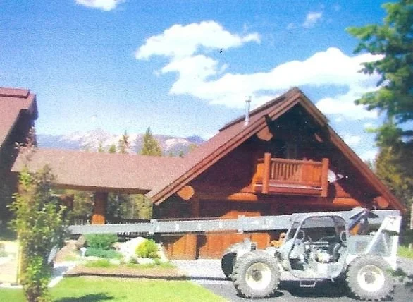 A wooden cabin with a sloped roof and a small balcony, with a golf cart parked in front, in a lush green area with trees and mountains in the background.