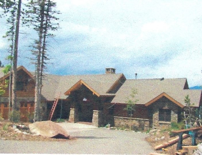 A large house with a wooden exterior and a stone foundation, situated in a wooded area with tall trees under a partly cloudy sky.