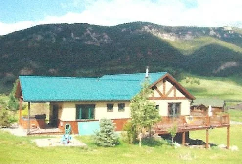 A house with a turquoise roof, beige walls, and a large wooden deck, surrounded by green grass and trees, with mountains in the background.