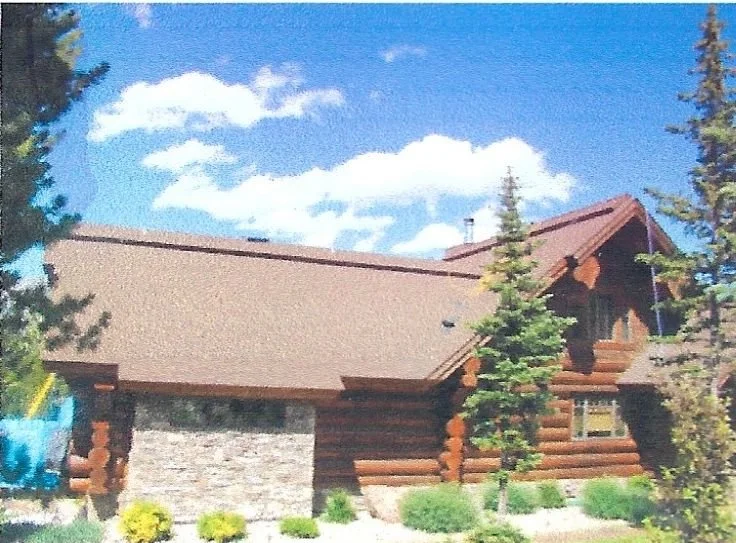 A log cabin with a sloped roof, surrounded by trees and shrubs under a partly cloudy sky.