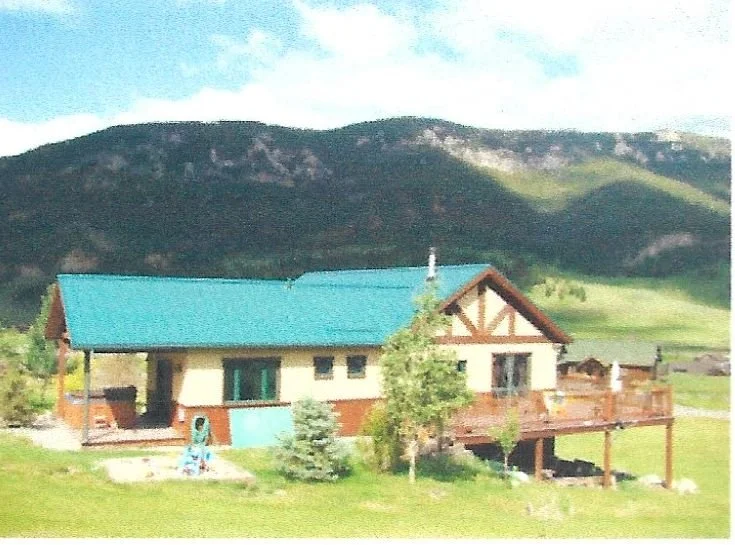 A house with a blue roof and wraparound porch, surrounded by green lawn and trees, with mountains in the background.