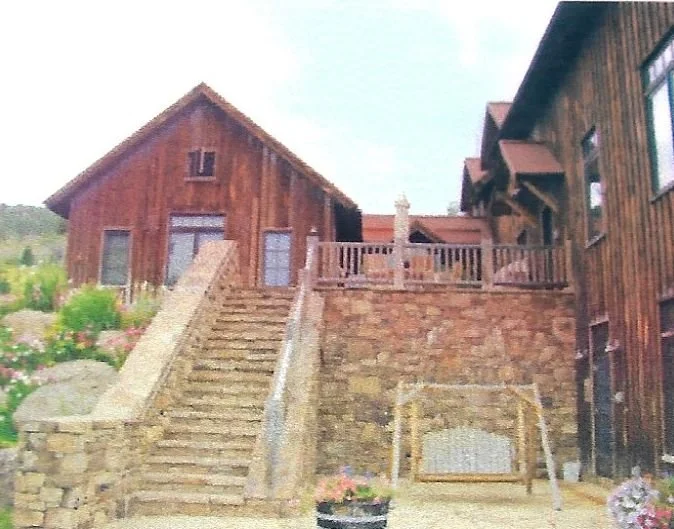 Exterior of a rustic wooden house with stone stairs leading up to a deck, surrounded by rocks and blooming flowers.