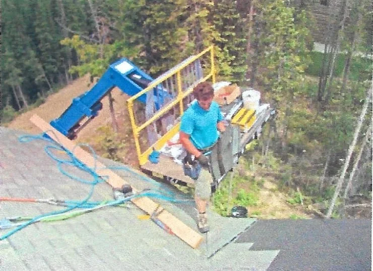 Worker standing on roof near a construction platform, surrounded by tools and equipment, with trees in the background.