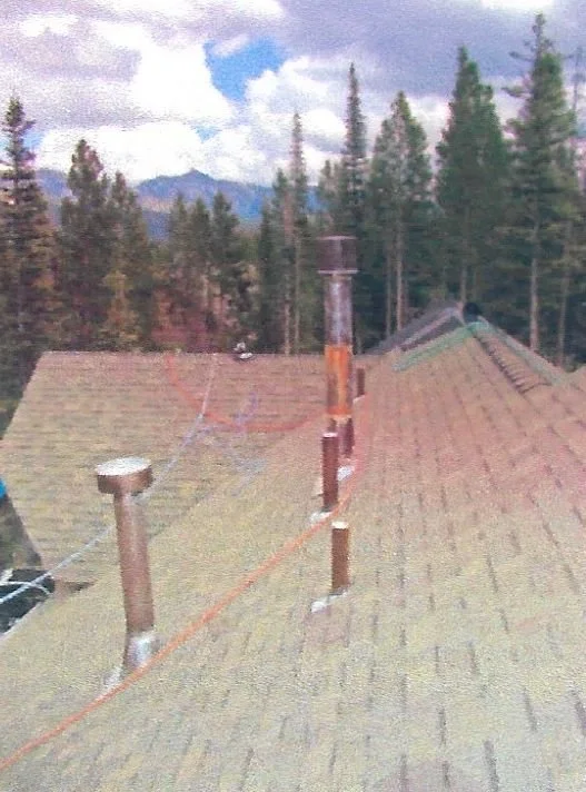 View of a rooftop with multiple metal chimneys and a safety wire, surrounded by pine trees and mountains in the background under a partly cloudy sky.