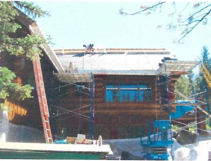 A house under construction with scaffolding and a ladder, partially covered by trees, and workers on the roof.