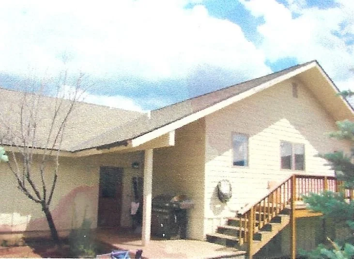 A beige house with a sloped roof, a small porch with a couple of windows, a wooden staircase leading to a door, a tree on the left side, and a grill below the staircase. The background shows partly cloudy skies.