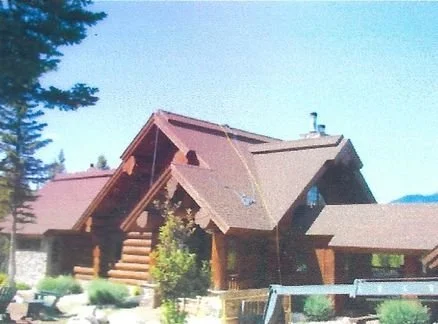 A log cabin with a steep roof and a chimney, surrounded by trees and greenery.