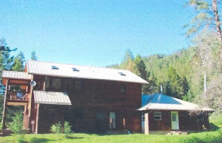 A two-story wooden house with a metal roof, surrounded by green grass and trees, with a smaller structure nearby, under a clear blue sky.