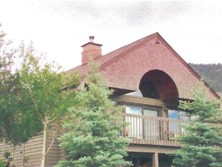 A two-story house with a red brick roof and chimney, surrounded by green trees, with a wooden balcony on the second floor, under a cloudy sky.