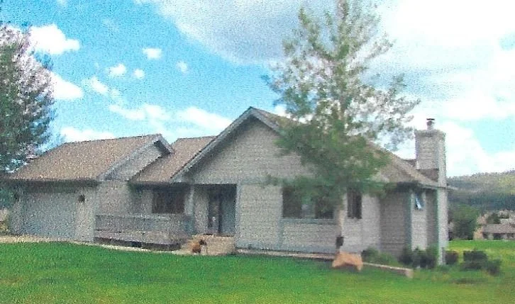 A house with gray siding, a porch, and a chimney, surrounded by a green lawn with trees against a blue sky with white clouds.