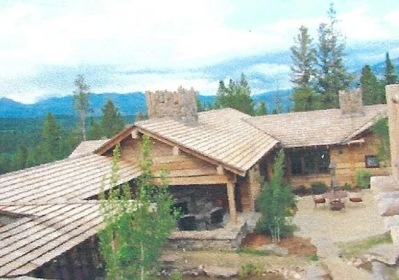 A wooden house in a forested area with mountains in the background, featuring outdoor seating and string lights.