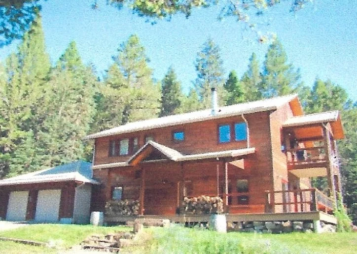 A multi-story wooden house with a deck, surrounded by trees, with a green lawn in front.