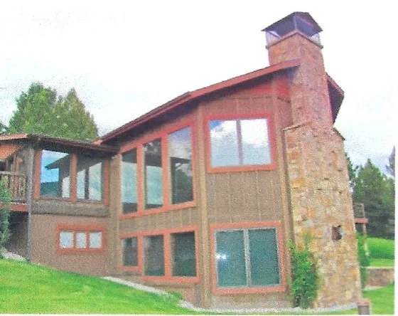 A two-story house with large front windows, a stone chimney, and wood siding, situated on a grassy hill.
