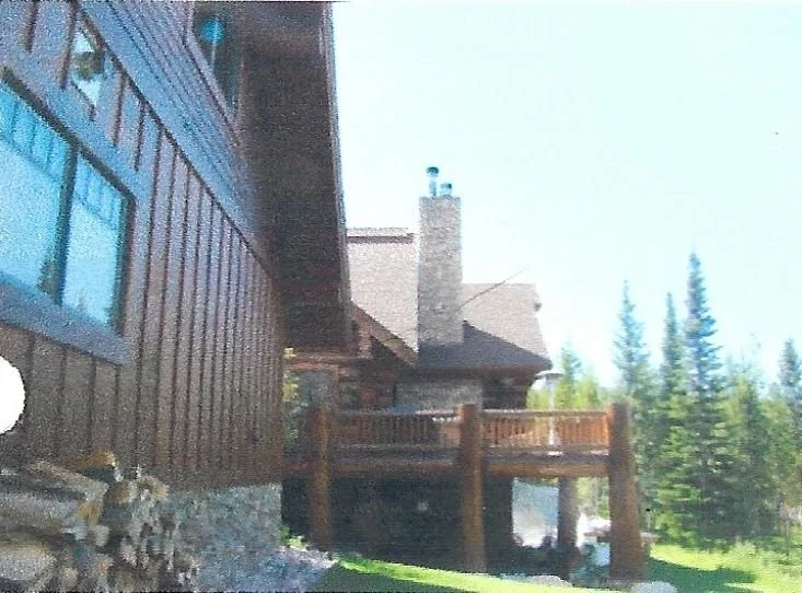View of a house with a dark wooden exterior and a stone foundation, a brown wooden balcony, and a chimney, with pine trees in the background.