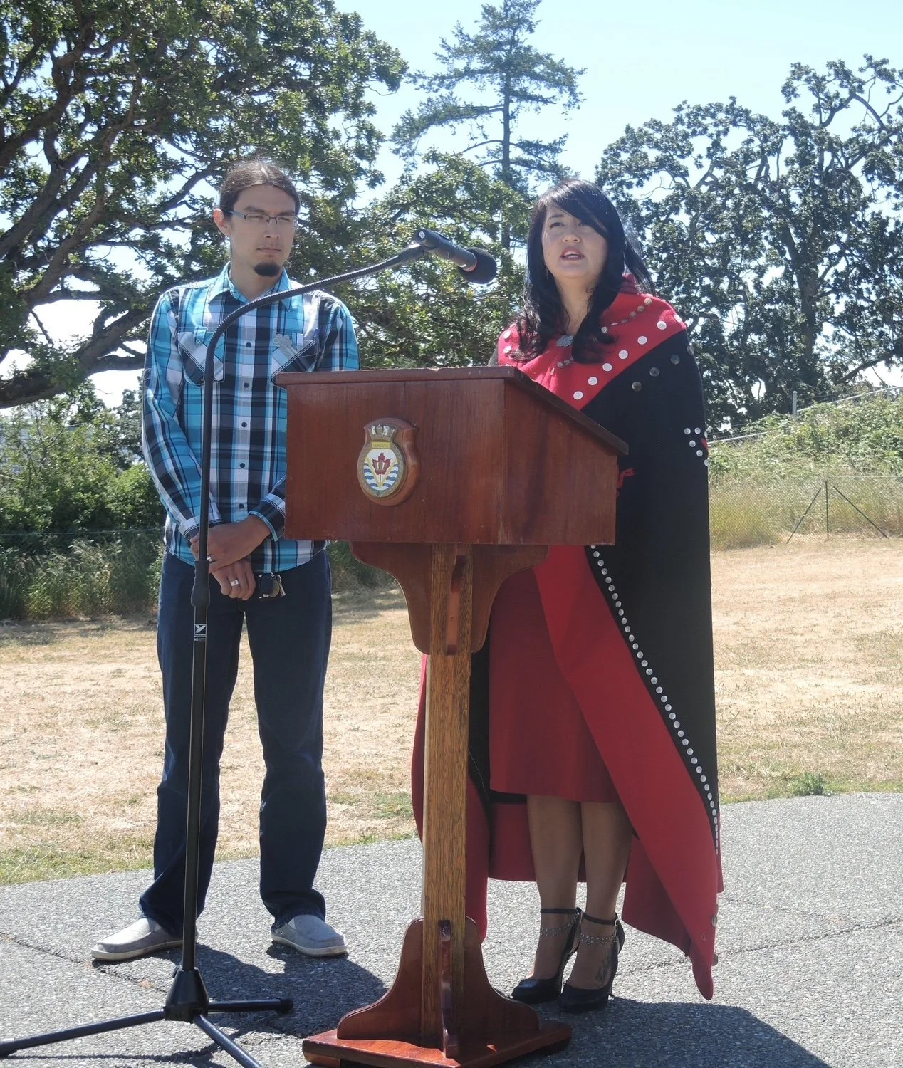 A woman in a red and black dress speaking at a podium outdoors, with a man in a blue plaid shirt standing beside her, and trees in the background.