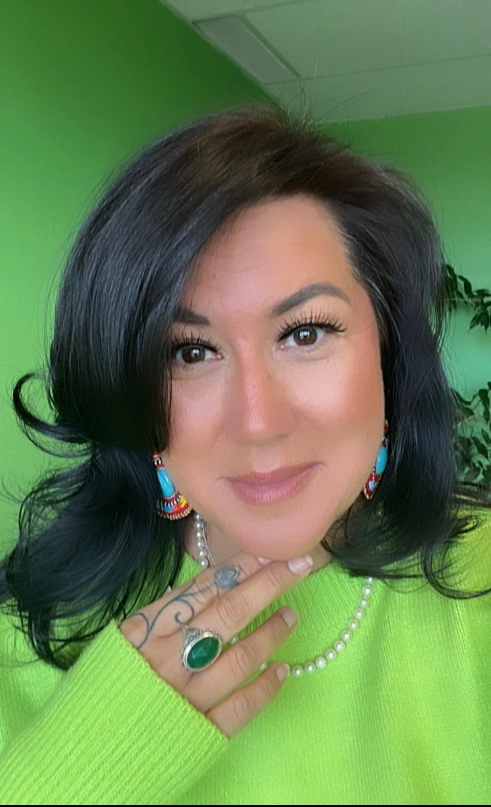 A woman with black, wavy hair, wearing a bright green sweater, turquoise earrings, a pearl necklace, and a large green ring on her finger, posing with her hand near her chin against a green background.