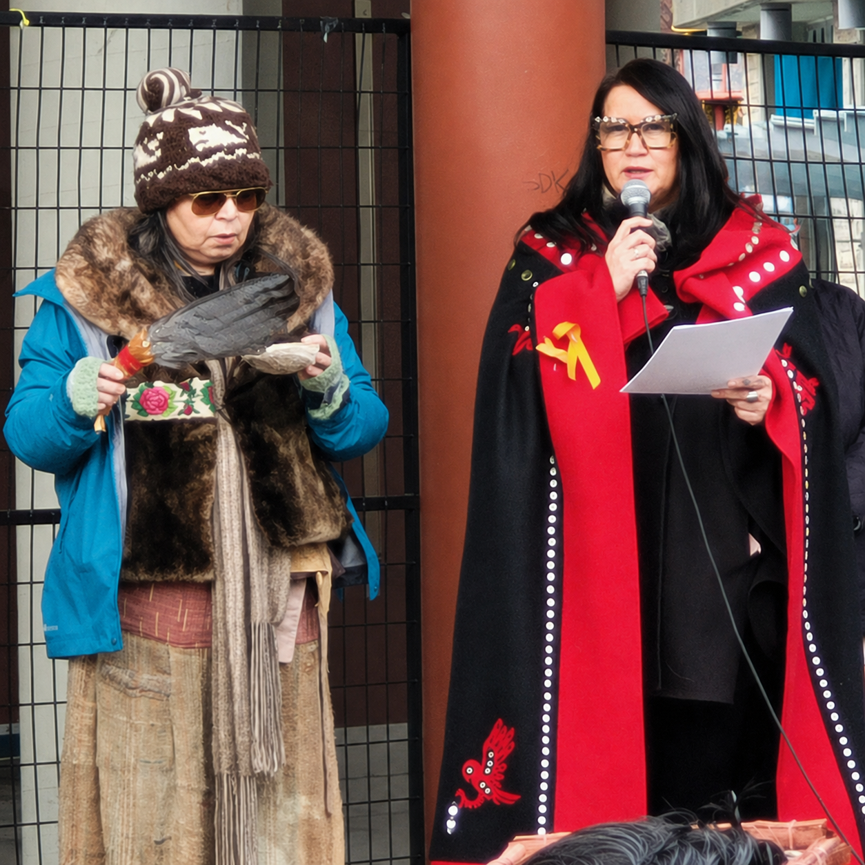 Two women standing outdoors against a black metal fence. The woman on the left is wearing a blue jacket, a brown and white knit hat, sunglasses, and is holding a large fish and a small container. The woman on the right is speaking into a microphone, 