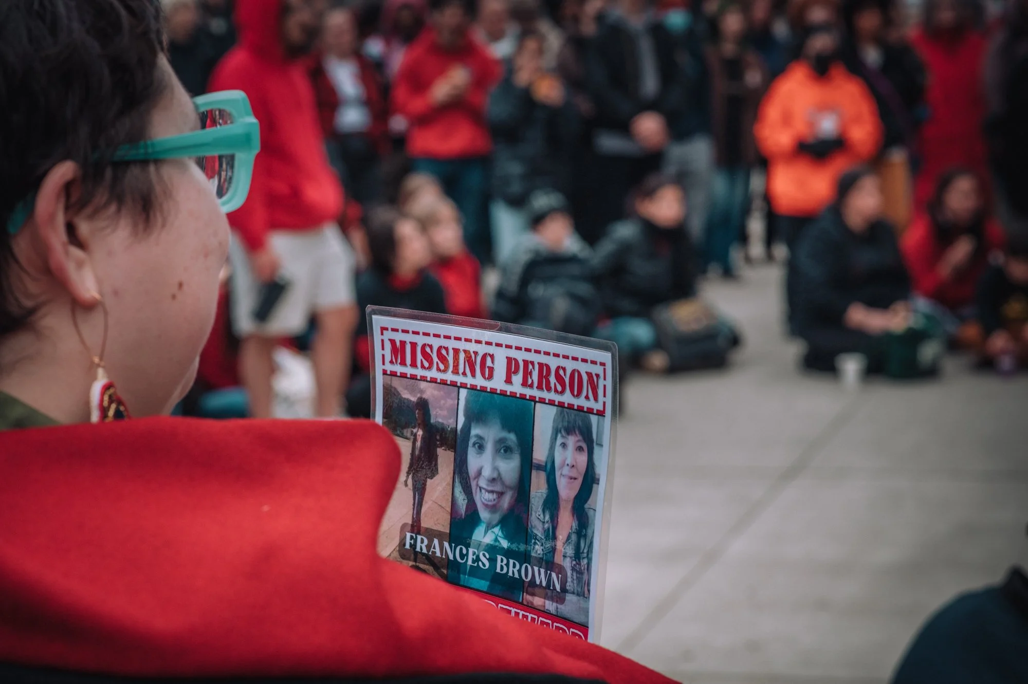 Close-up of a woman with short dark hair and earrings, wearing turquoise glasses, holding a missing person poster with photos of a woman named Frances Brown. The background shows a crowd of people seated, dressed in dark or red jackets, at an outdoor