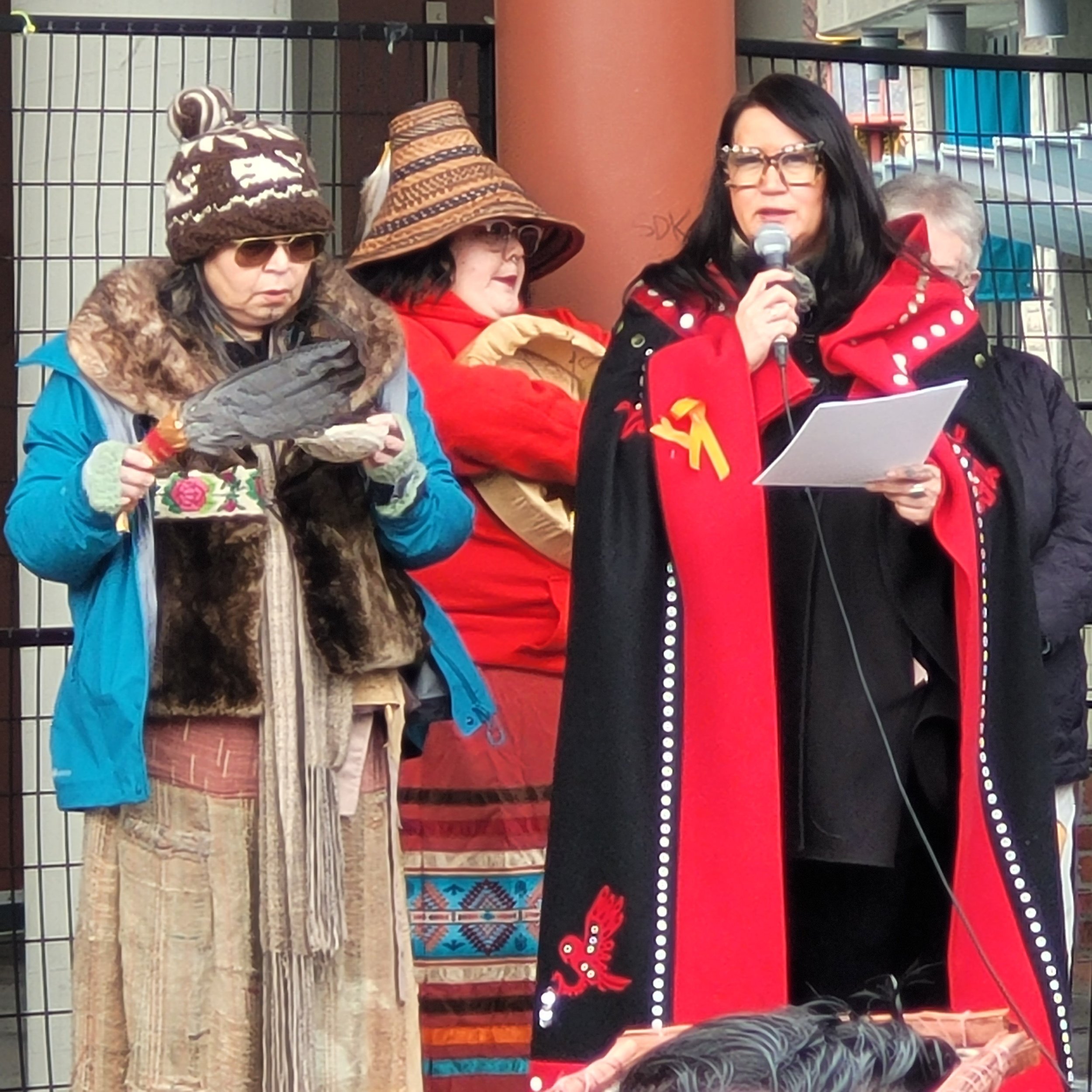 A woman with black hair and glasses speaking into a microphone during an outdoor event, wearing a red and black outfit with white polka dots, surrounded by older women in warm clothing and hats.