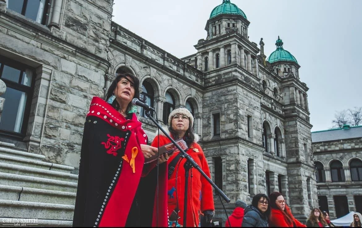 Two women speaking at a podium outside a historic stone government building, with people sitting on steps watching.