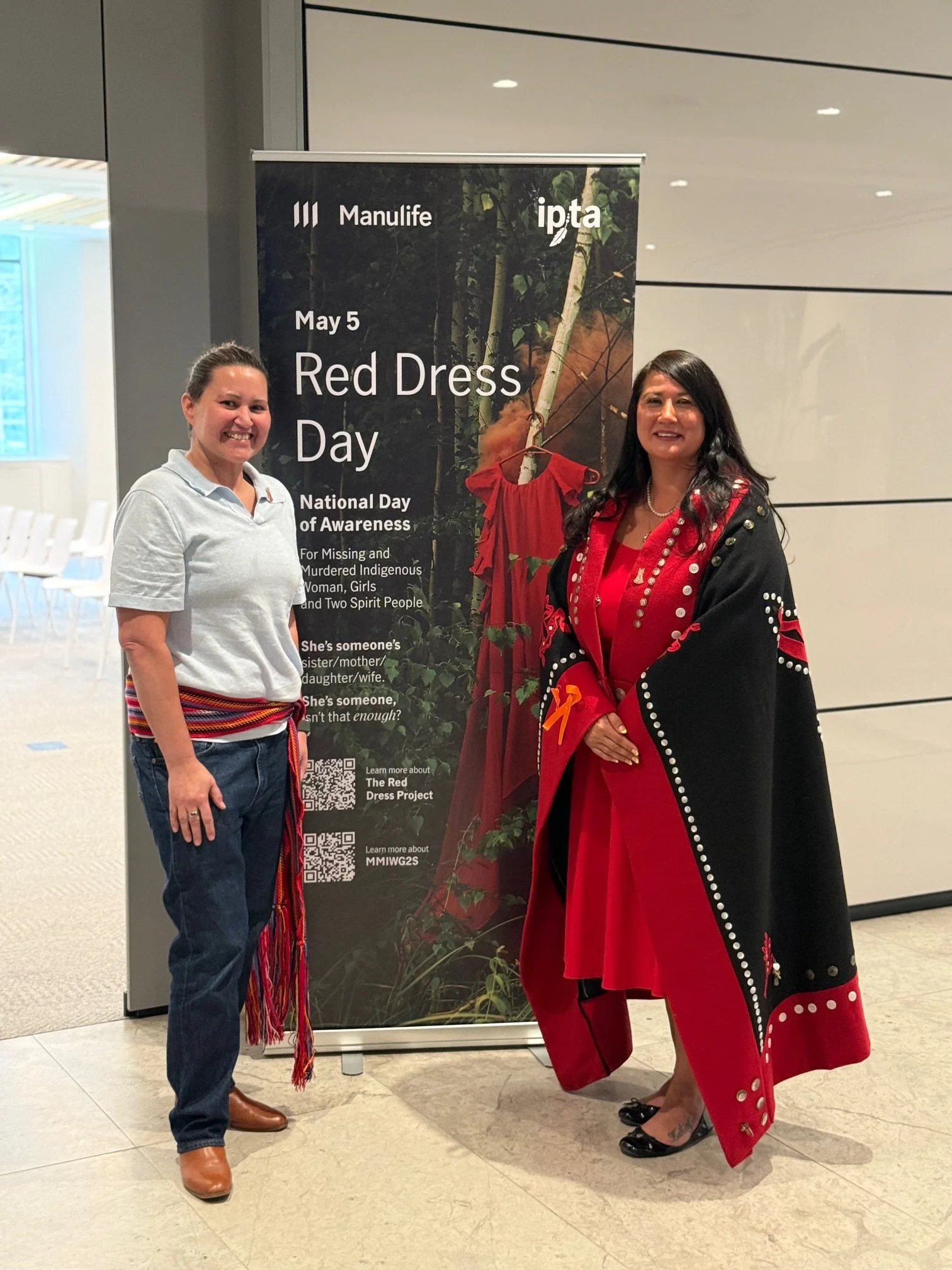 Two women standing in front of a vertical banner promoting Red Dress Day on May 5th, part of National Day of Awareness, with a background of chairs and a modern interior.