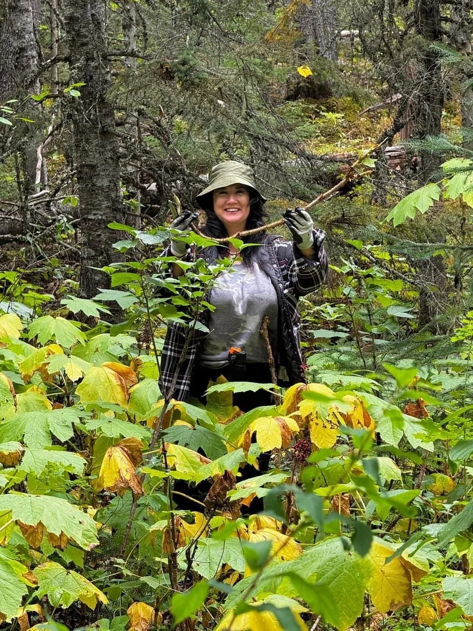 A woman smiling and holding a stick, standing in a dense forest surrounded by green and yellow foliage.