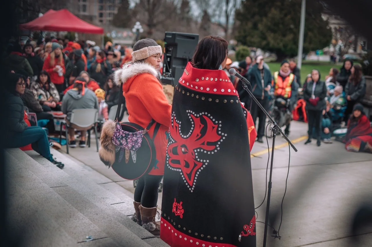 Two women speaking into microphones at a public outdoor event with a large crowd gathered behind them, some sitting and some standing, in a park-like setting.