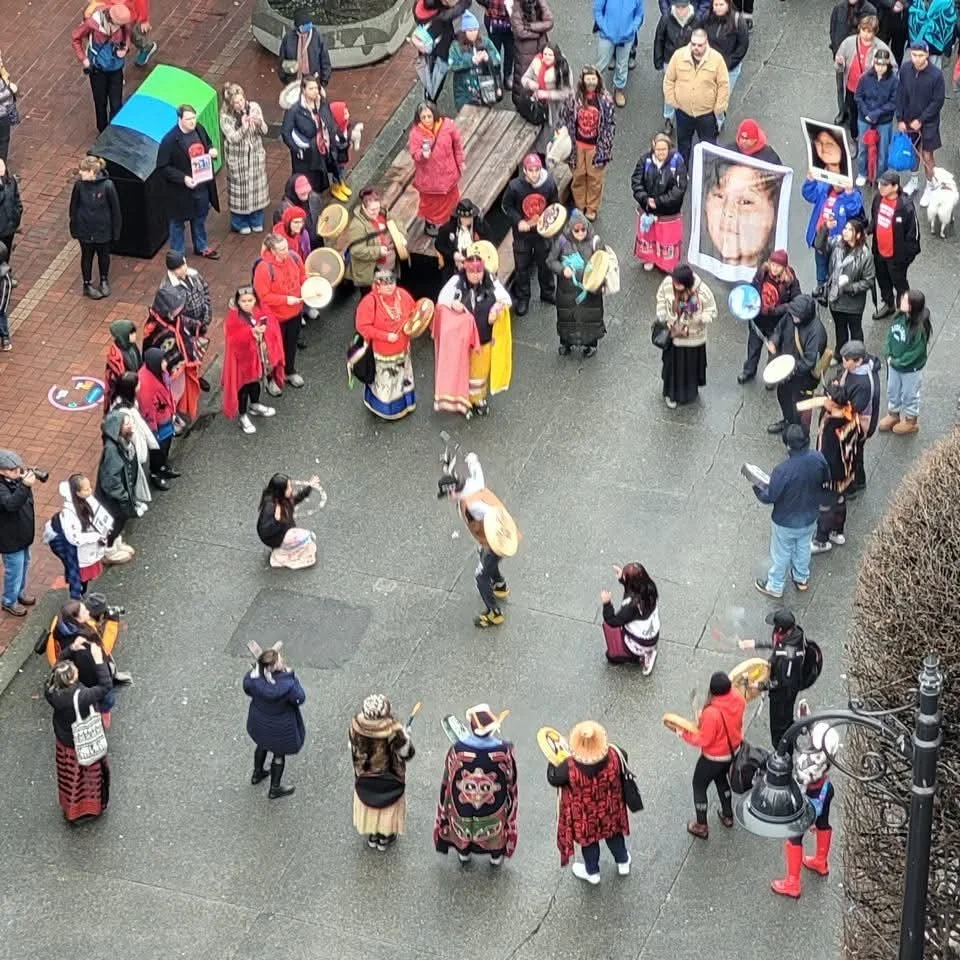 Group of people gathered in a circle, some wearing traditional clothing and hats, playing drums, and others taking photos or watching. Large photos and banners are displayed among the crowd, with some individuals holding signs and camera equipment ca