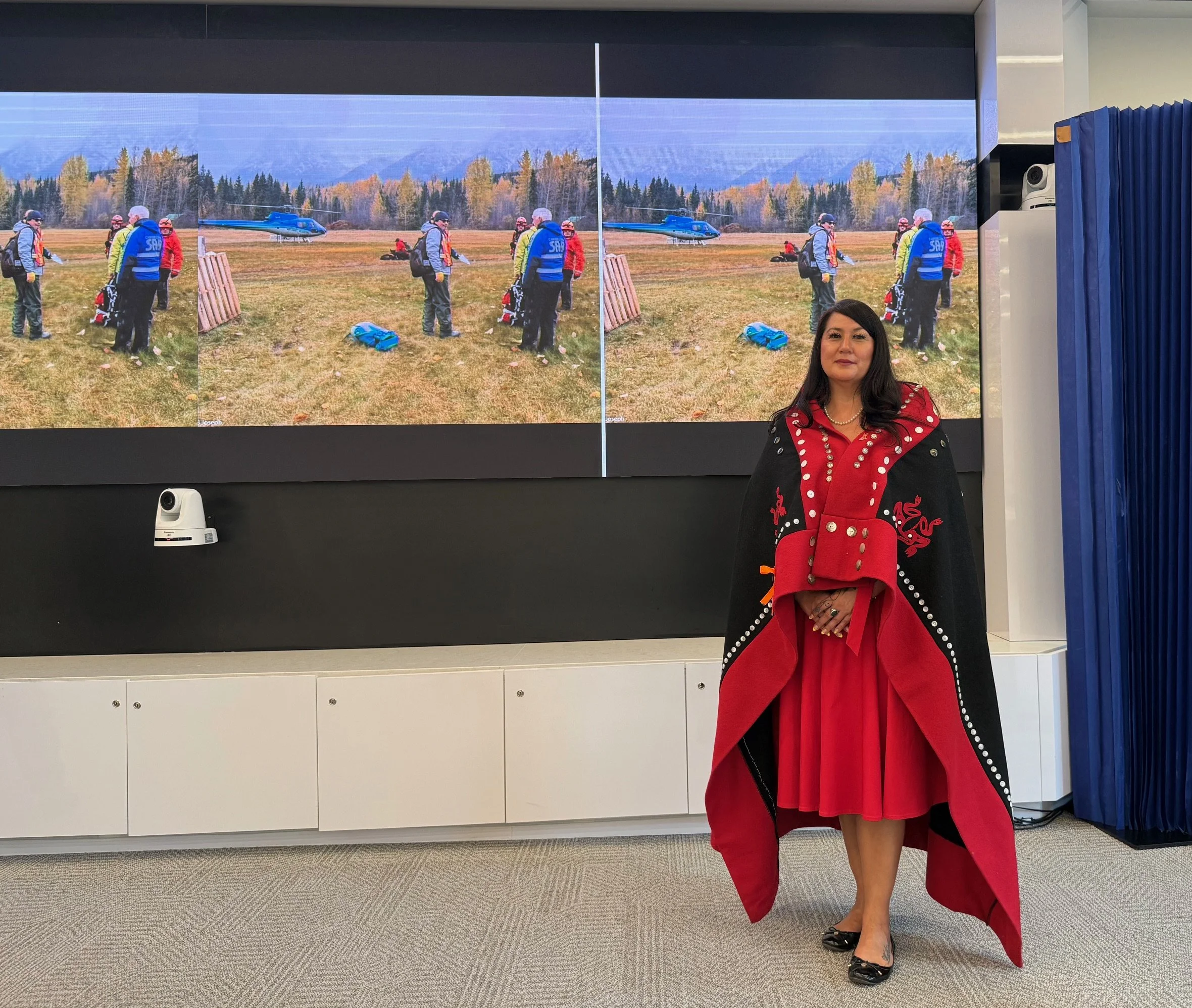Woman with long dark hair wearing a red and black jacket with decorative pearl buttons, and a matching red skirt, standing in front of a large digital screen displaying an outdoor scene with people and helicopters. She is smiling and wearing high hee