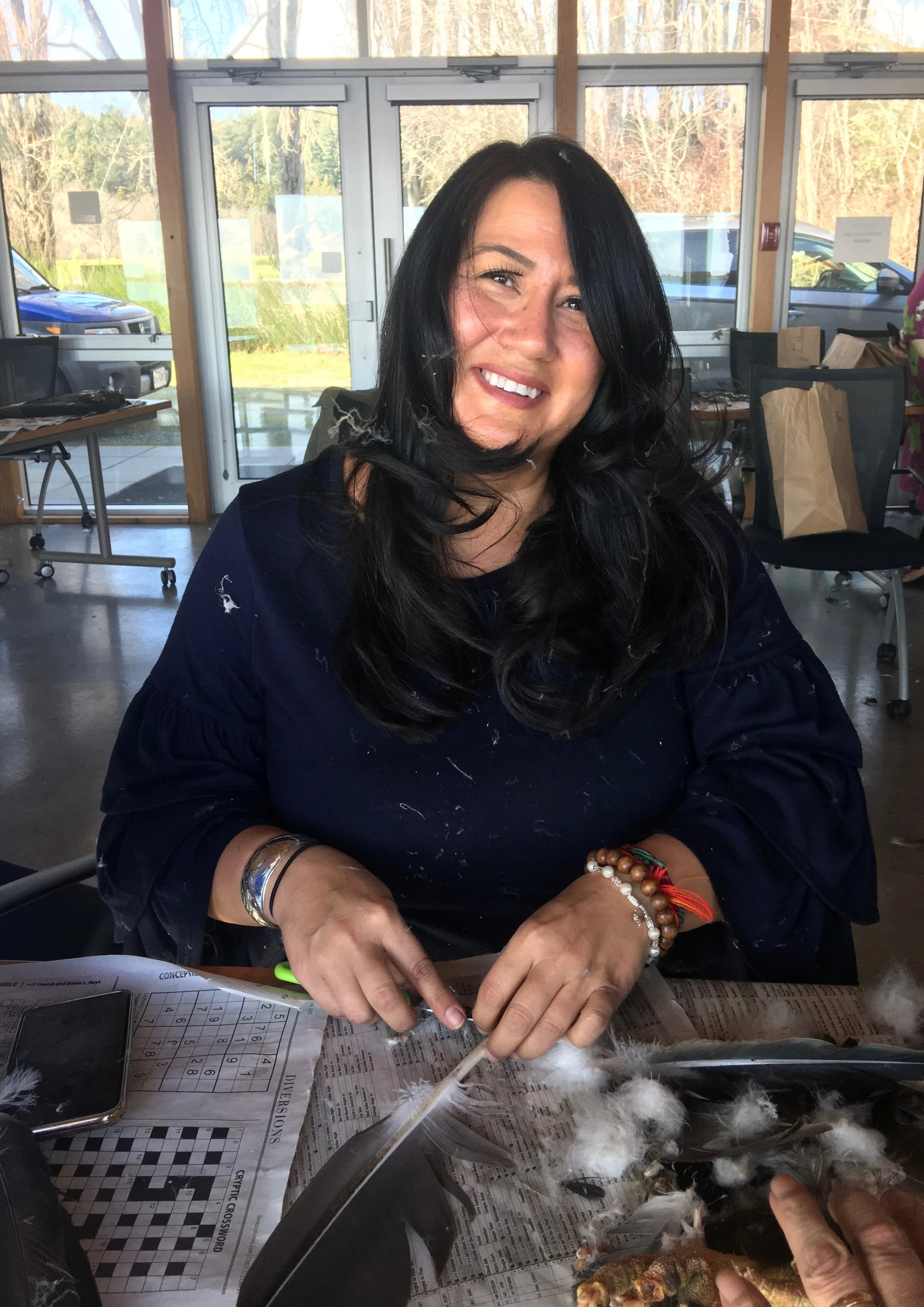 A woman with long black hair smiling at a table filled with a newspaper, a crossword puzzle, a smartphone, feathers, and various objects. Behind her, large windows reveal parked cars and a wooded outdoor area.