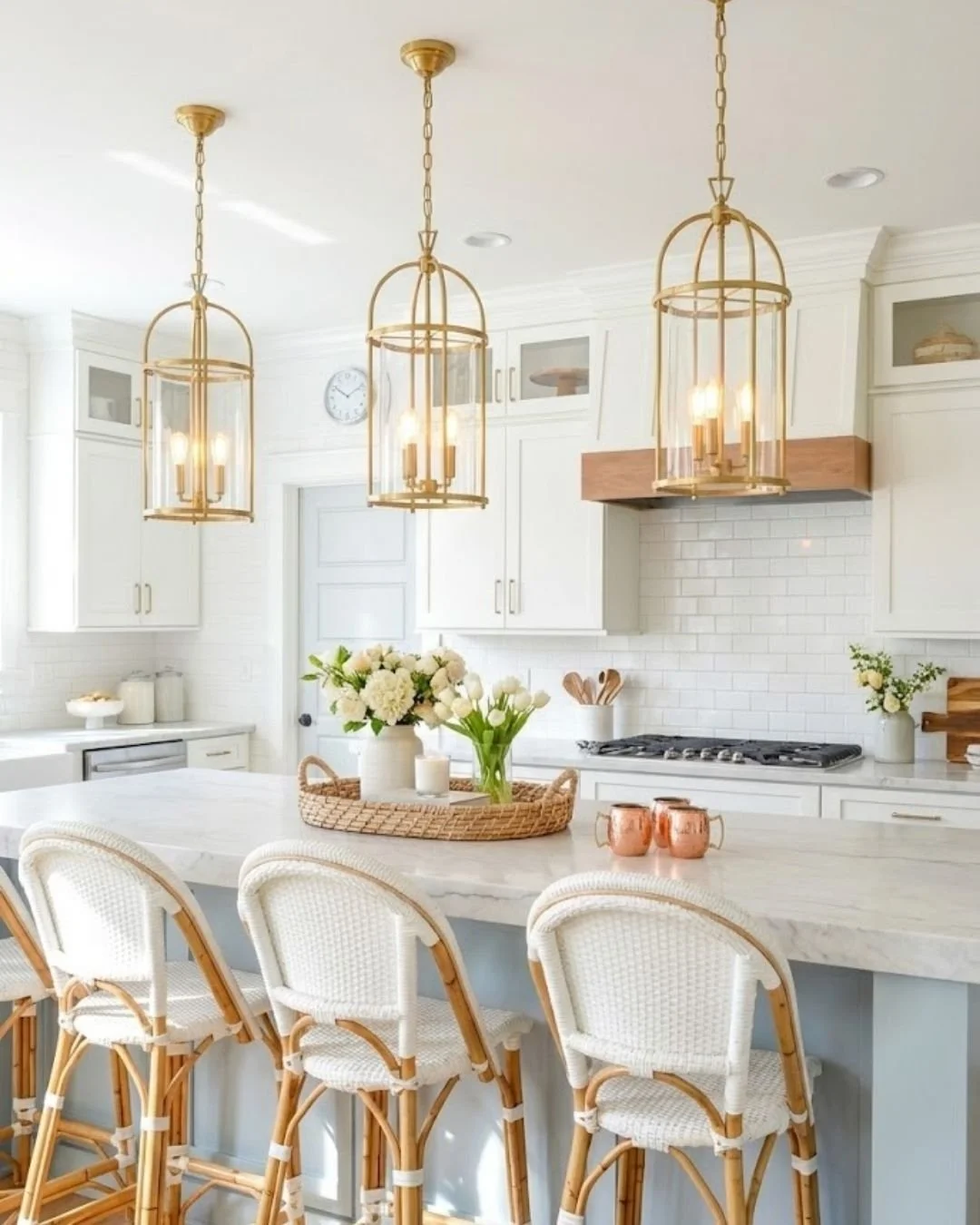 A bright, modern kitchen with white cabinets, a white marble island, three gold pendant lights, floral arrangements, and tan wicker bar stools.