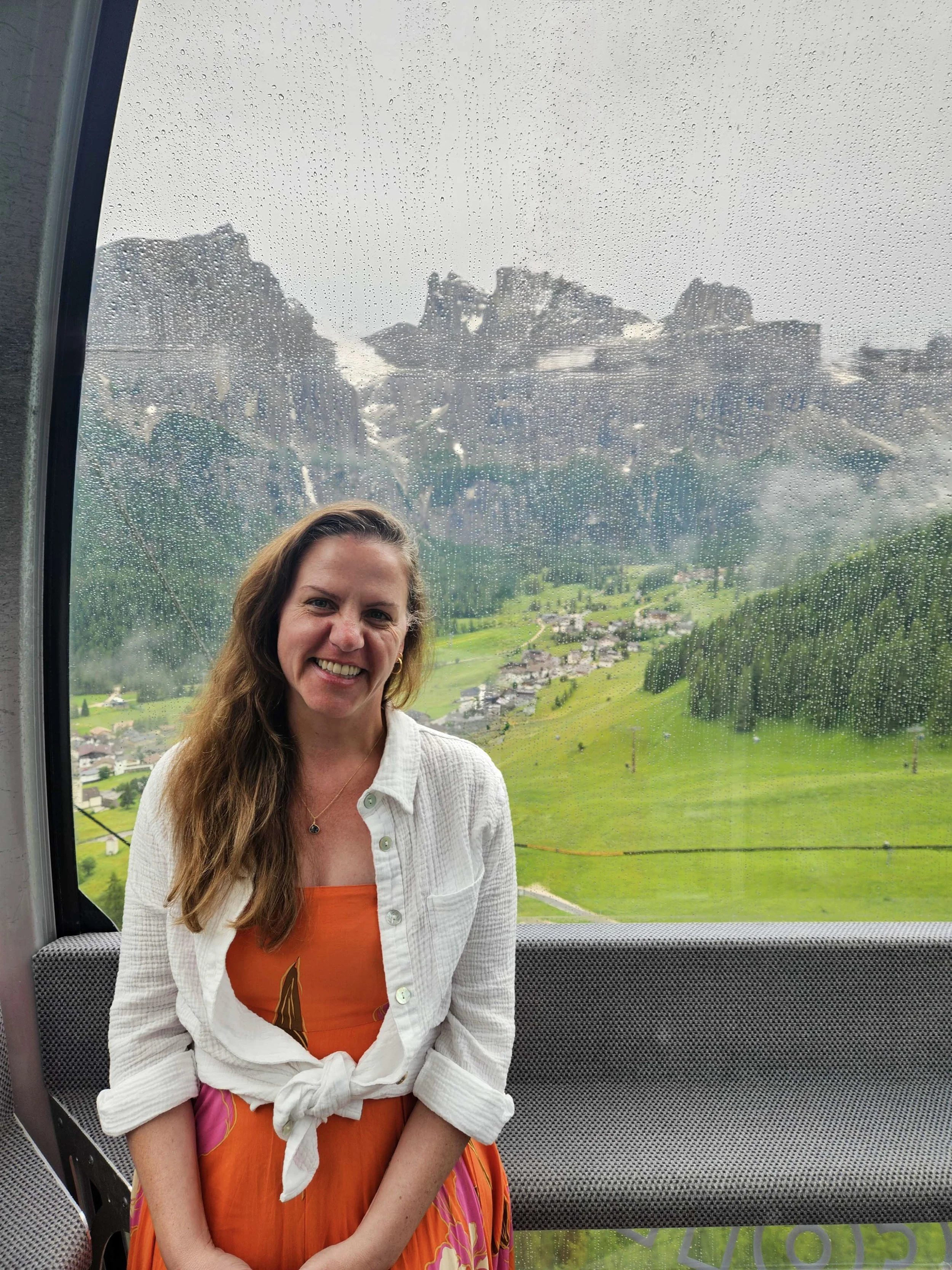 A woman smiling inside a cable car with sweeping views of green Dolomite valleys and rocky peaks in the rain, Italy