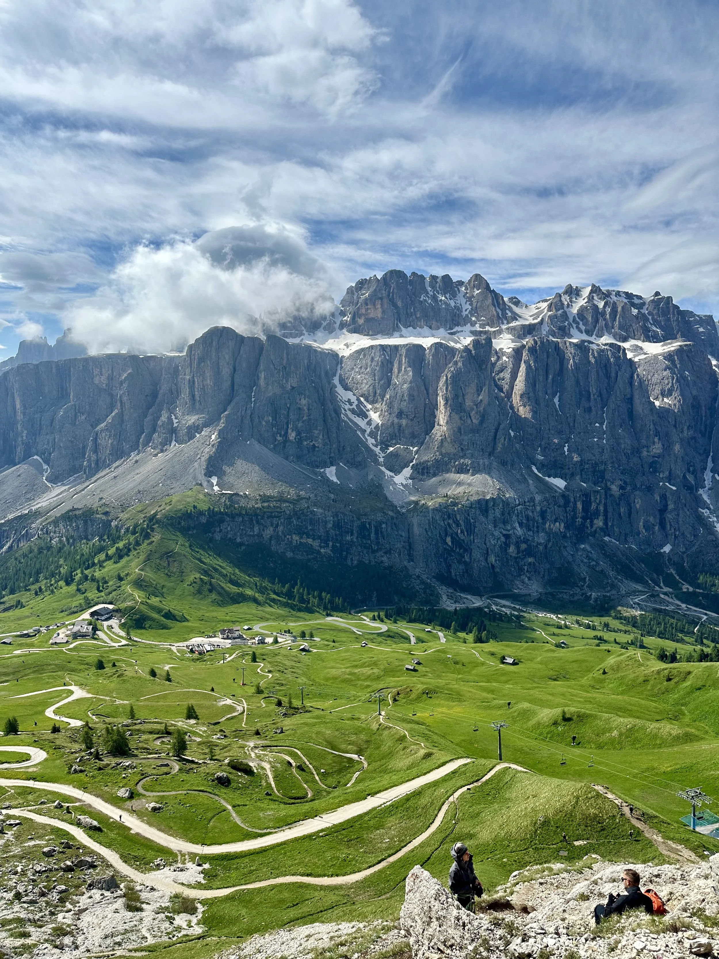 Sweeping panoramic view of jagged Dolomite peaks, green alpine meadows, and winding mountain trails in Italy