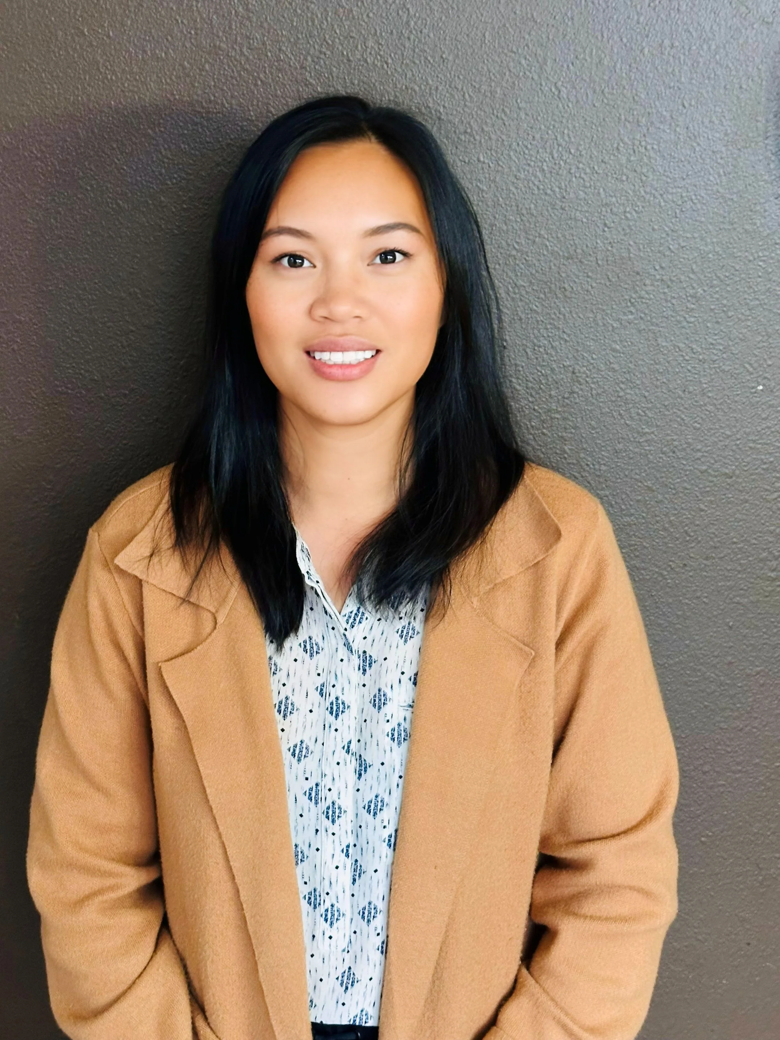 A woman with black hair, wearing a camel-colored coat, a white shirt with a blue pattern, and sitting against a gray textured wall.
