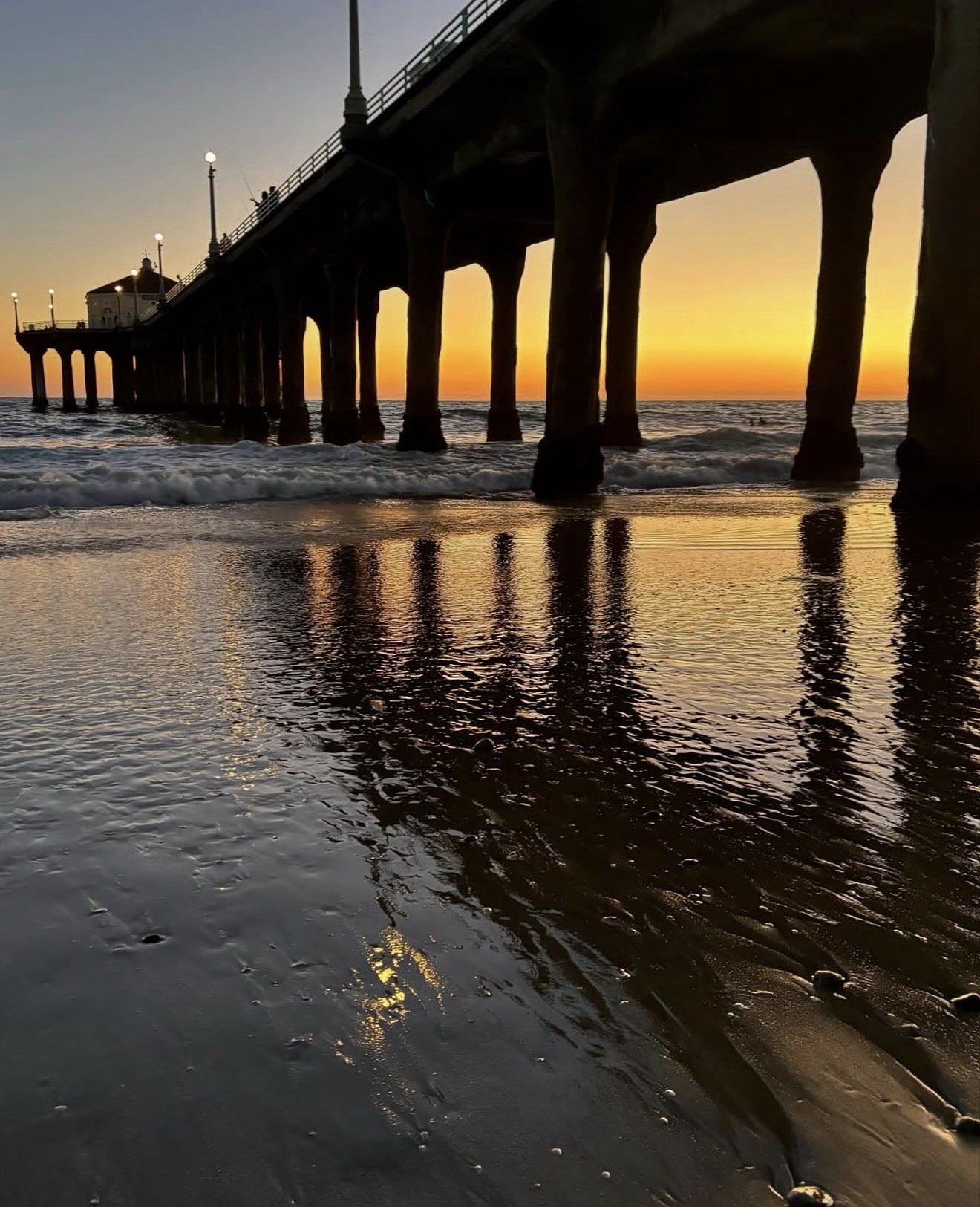 Sunset view at the beach with a pier extending over the ocean, reflecting warm colors on the wet sand, with gentle waves lapping at the shore.