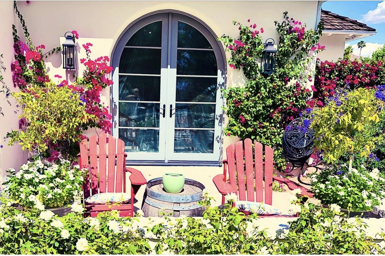 Faith- based life coaching, A colorful outdoor patio scene with two pink Adirondack chairs, surrounded by vibrant flowers and greenery, in front of a house with a large arched window and black lantern-style wall lights.