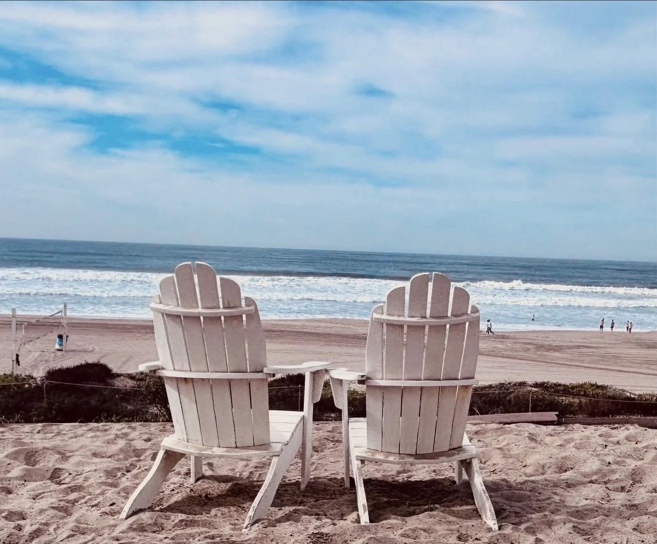 Two white Adirondack chairs on a sandy beach facing the ocean and a blue sky with some clouds above.