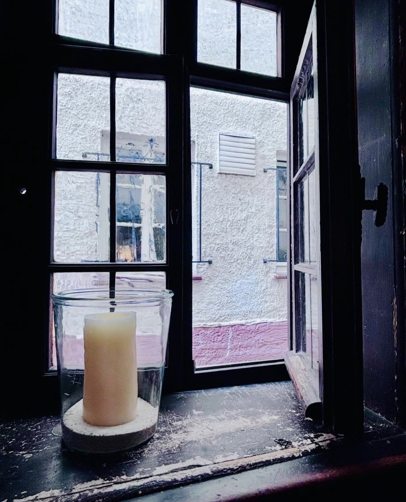View from inside a home looking out a window and door to a small courtyard with textured white walls and a red floor. There is a large candle in a glass holder on the window sill.