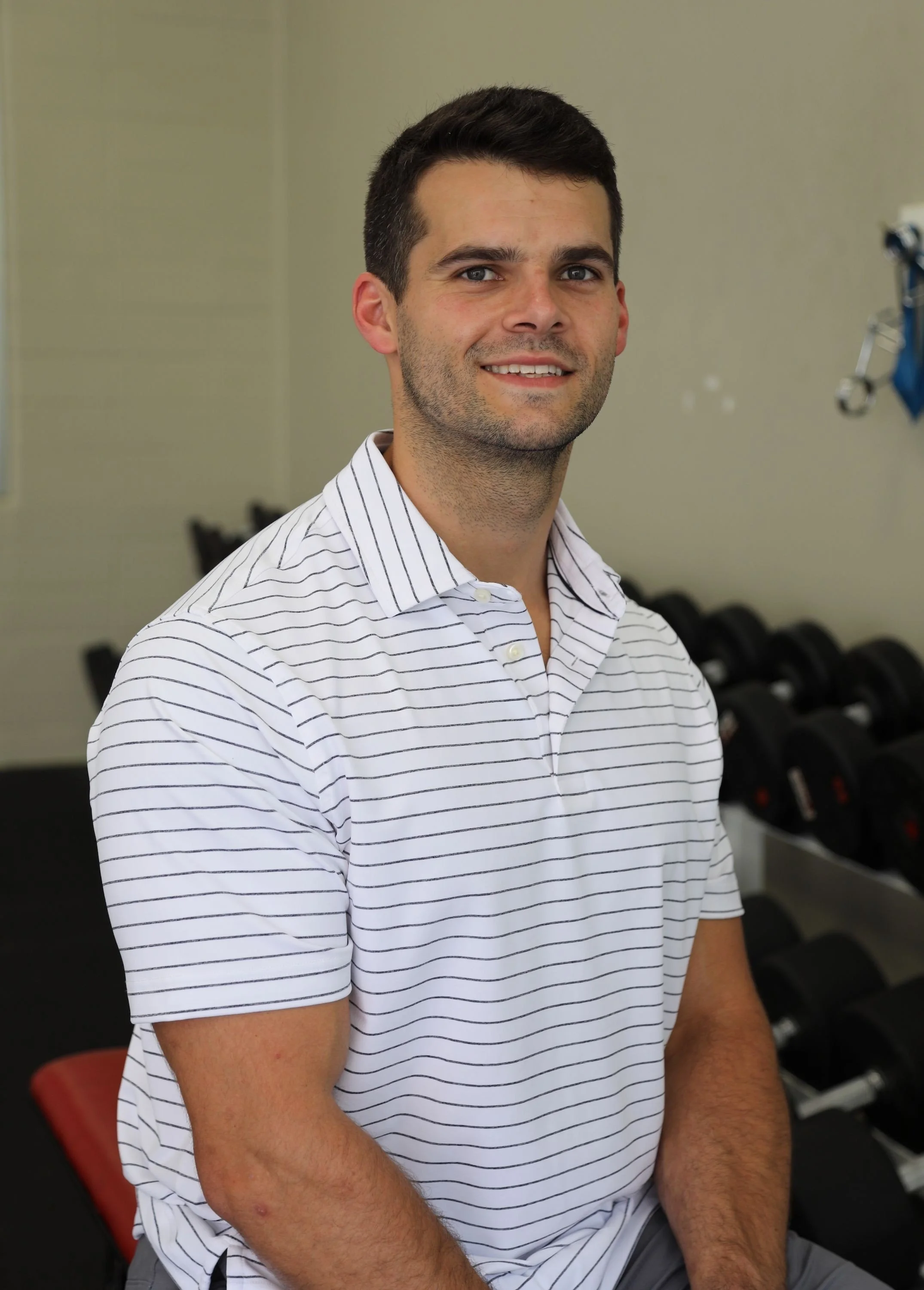 Young man with short dark hair, light beard, wearing a white and gray striped shirt, smiling, sitting in a room with black dumbbells in the background.