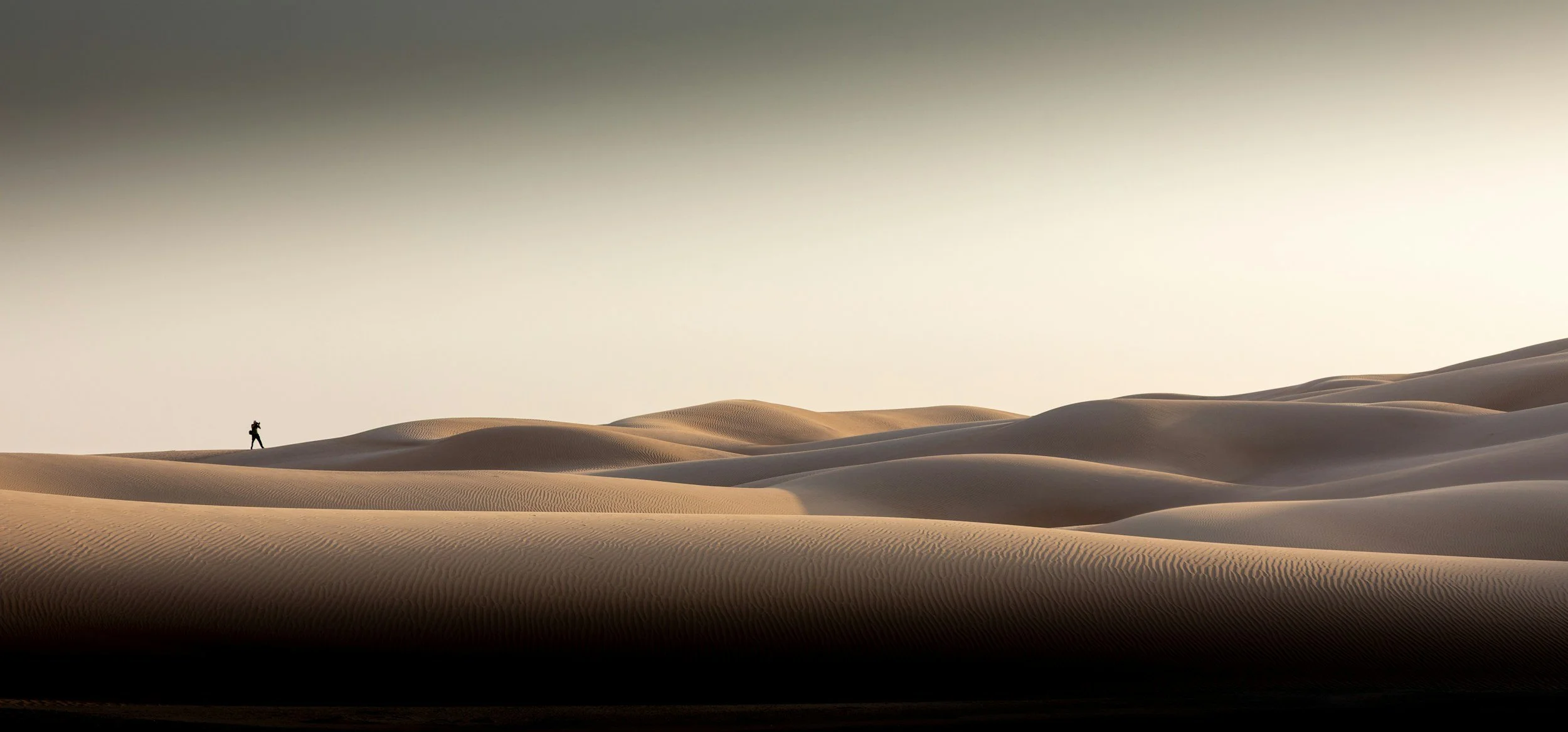 A solitary person walking across sand dunes in a desert landscape, with a vast open sky above.