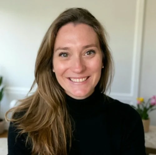 A woman with long, light brown hair smiling at the camera, wearing a black top in a room with a white wall and pink and yellow flowers in the background.