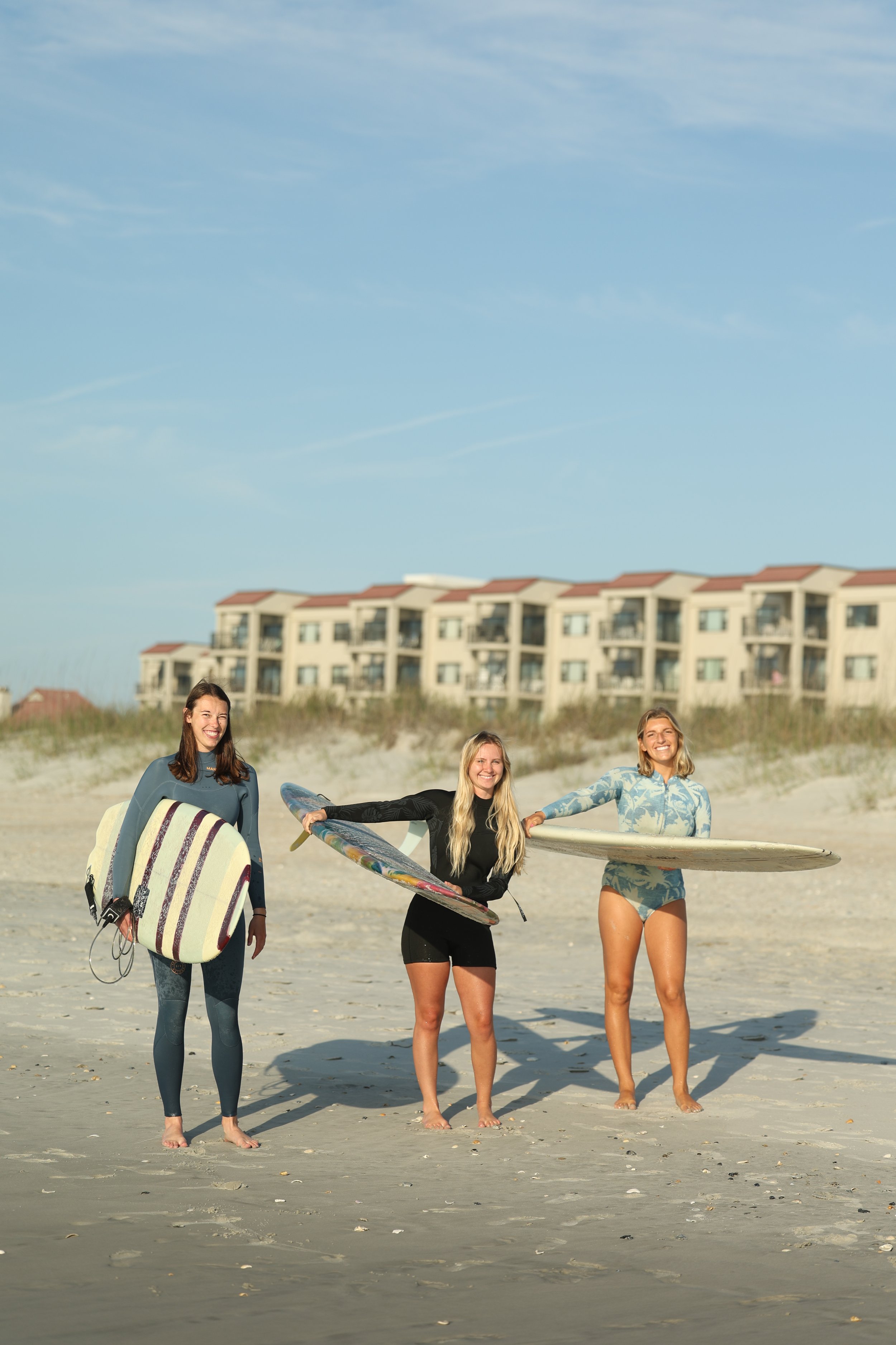 Four young women standing on the sand at the beach, holding surfboards, and smiling. In the background, there are beachside apartments and a blue sky with light clouds.
