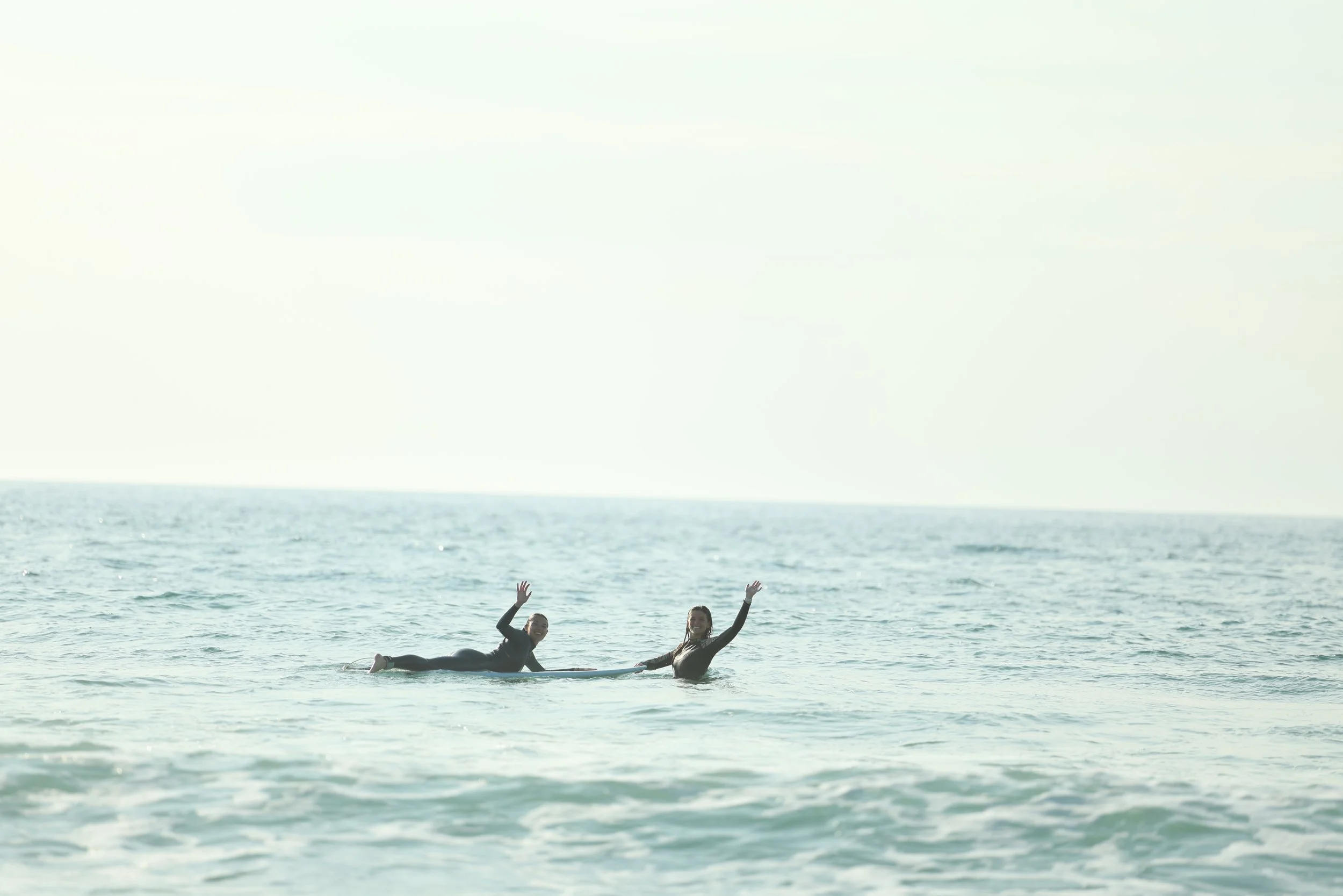 Two people in wetsuits are floating in the ocean, smiling and waving at the camera, with a clear sky above.