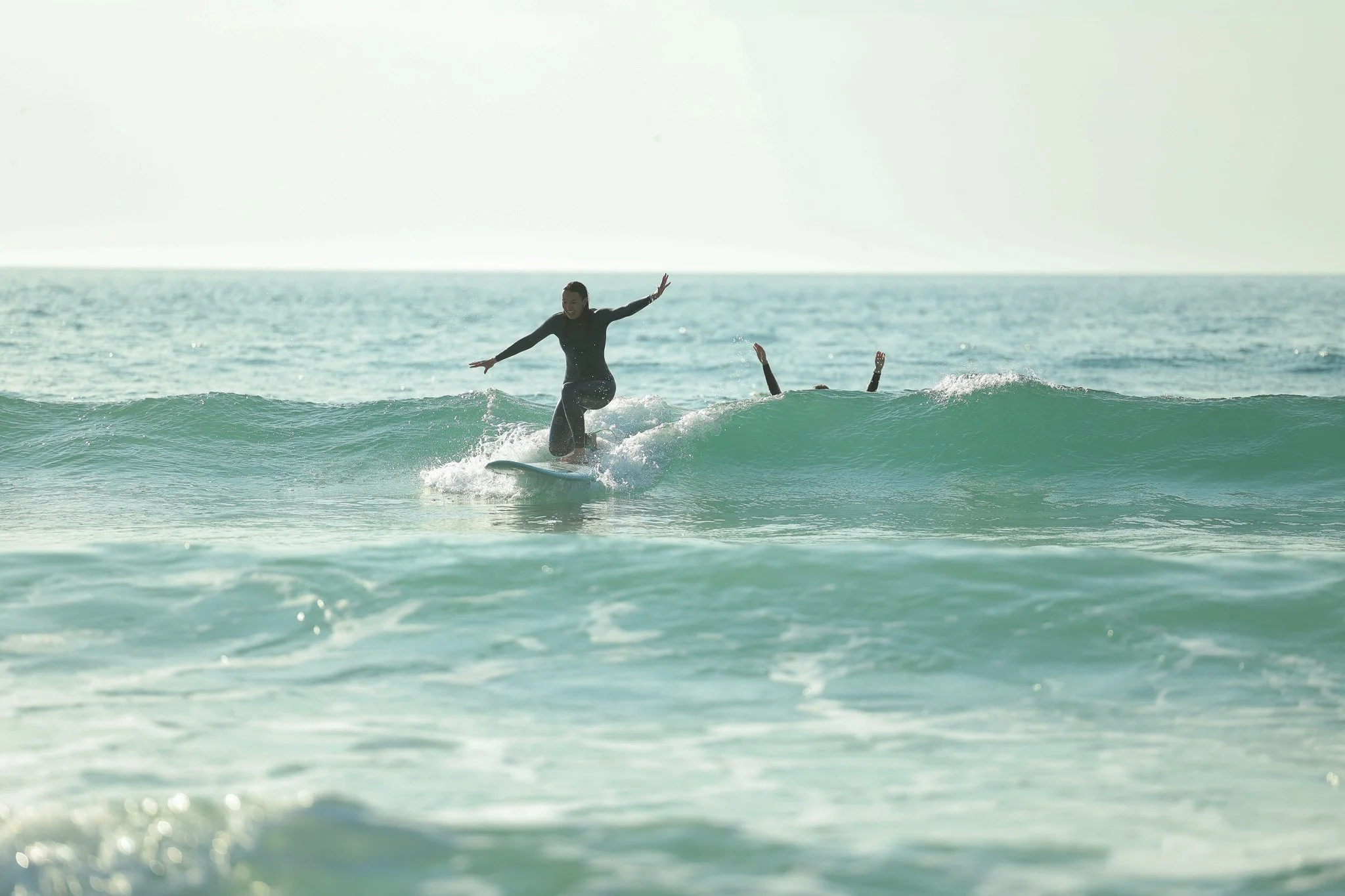 Person surfing on a wave in the ocean with another person cheering behind them, both wearing black wetsuits.