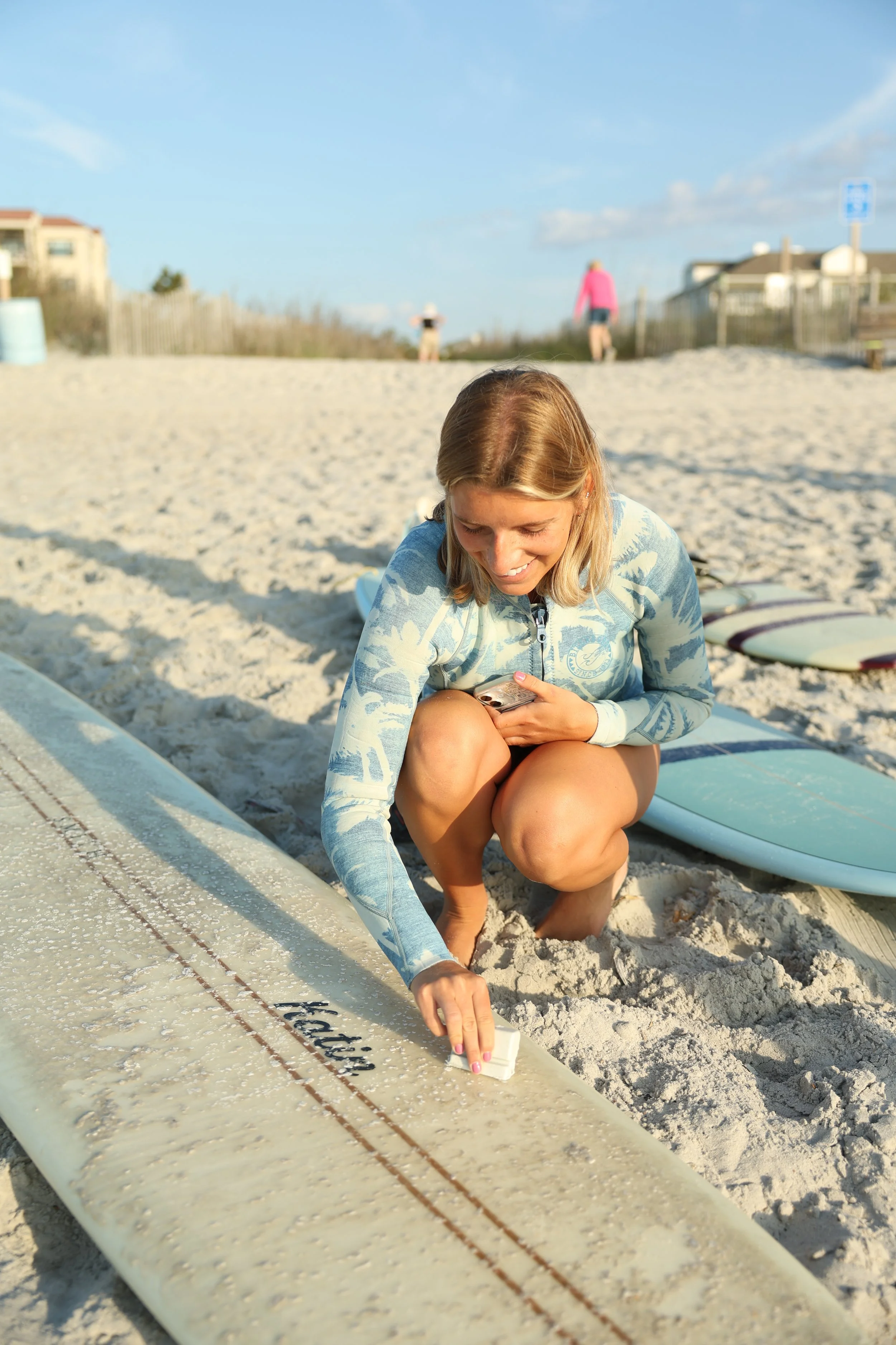 A woman in a blue and white patterned rash guard and shorts kneels on the sand, applying wax to her surfboard. In the background, other beachgoers are walking along the beach, with houses and a clear blue sky behind a fence.