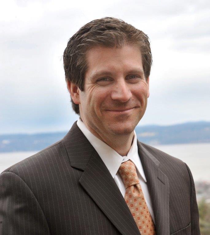 A smiling man in a dark suit and orange tie standing outdoors with water and mountains in the background.
