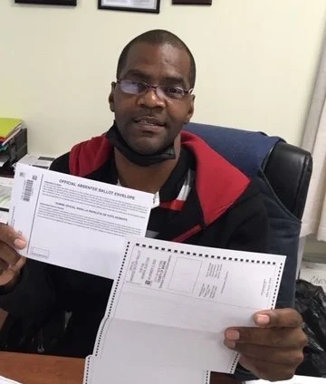 Man with glasses sitting at a desk holding two documents or forms.
