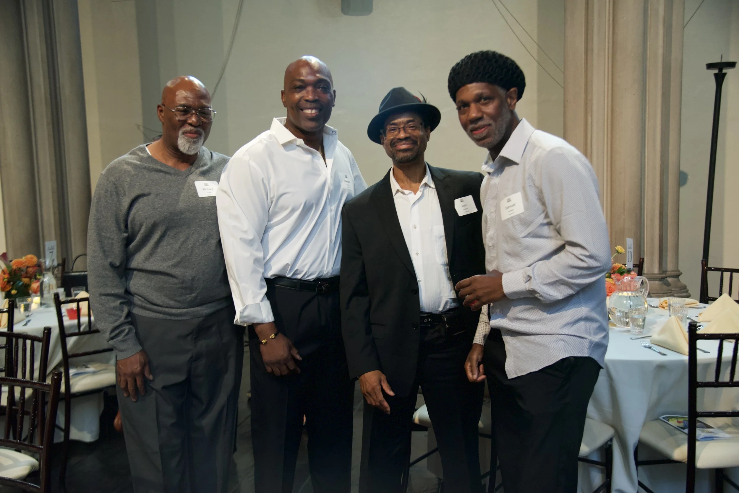 Five men are standing together in formal attire at a banquet or conference, smiling for the camera. They are in a decorated room with round tables set with tableware and napkins.