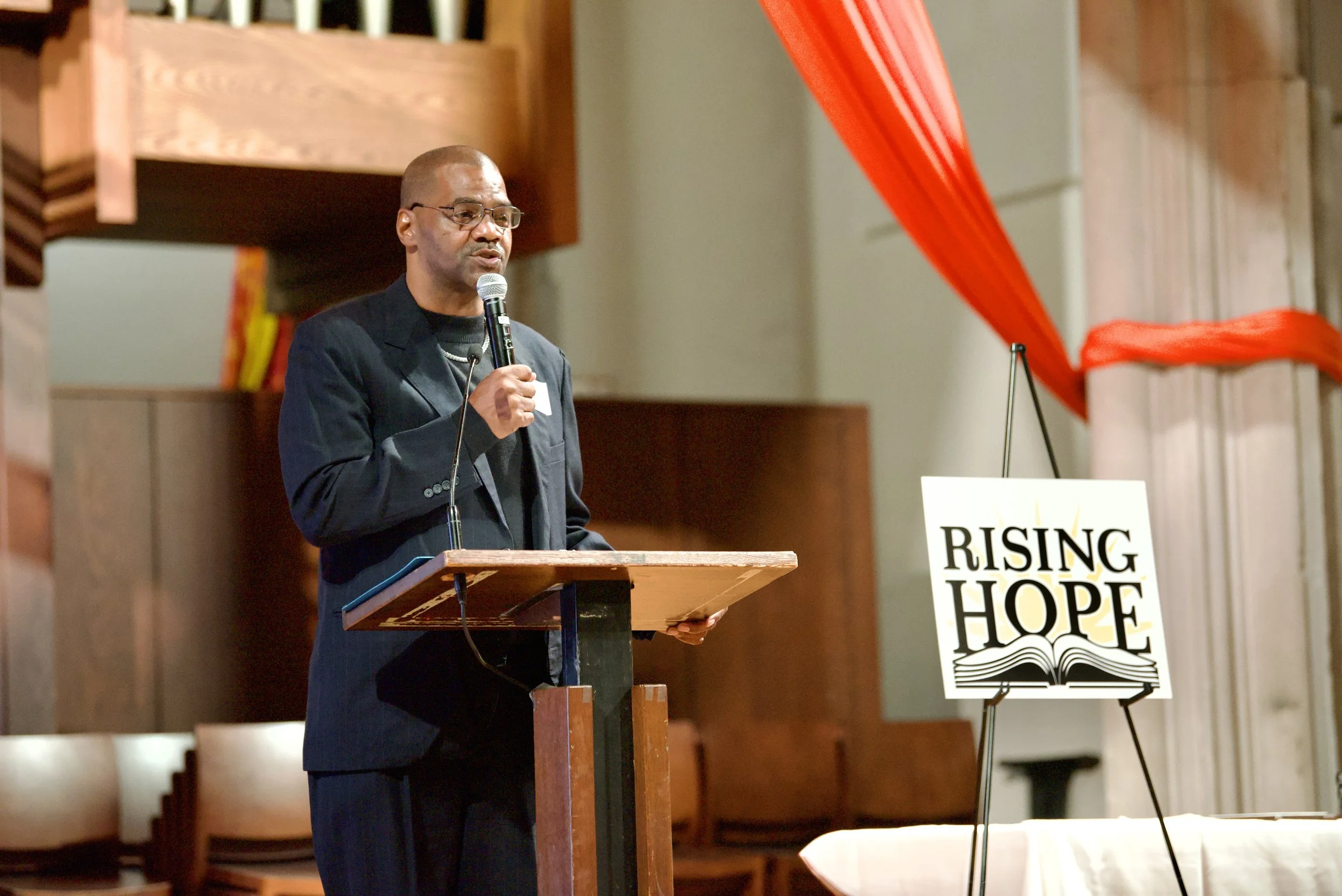 A man in glasses and a dark suit speaking into a microphone behind a wooden podium. There is a sign that reads 'RISING HOPE' with images of an open book and a bird on it. Red fabric is draped across poles to the right of the man.