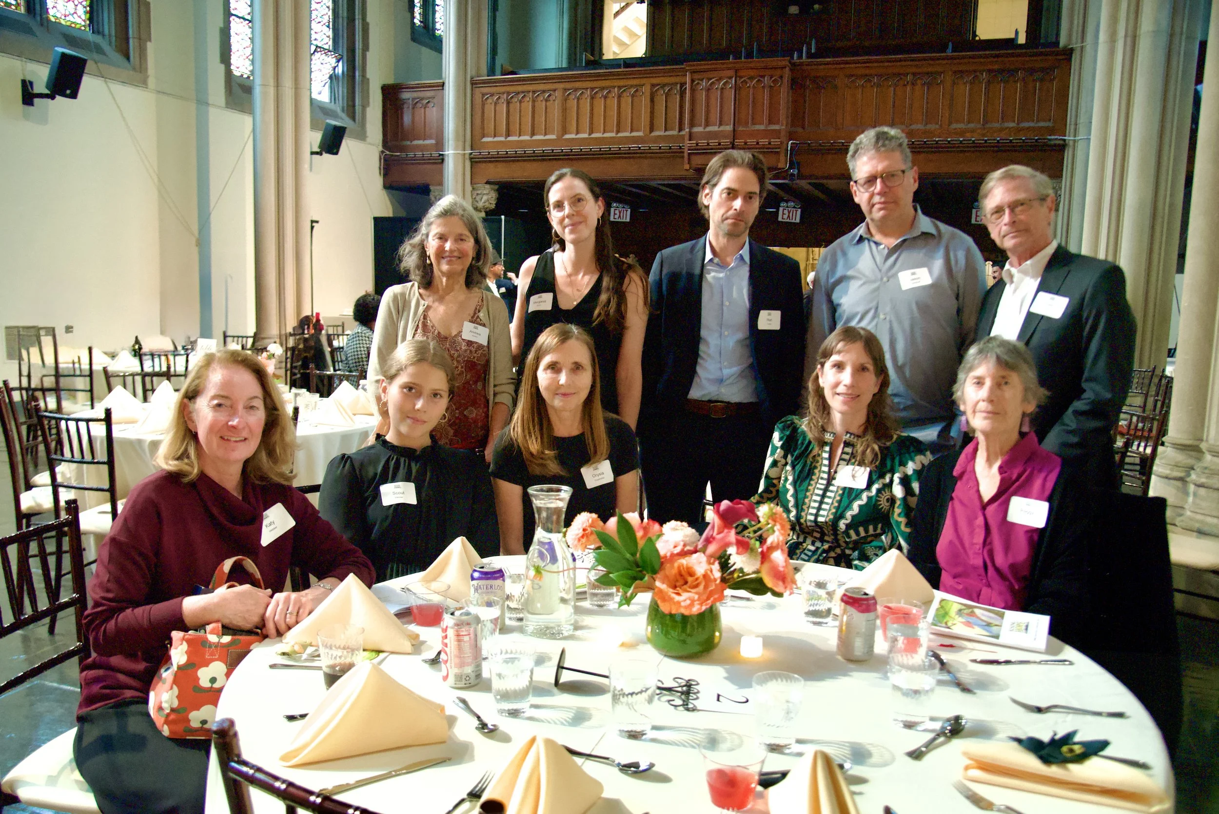 Group of people at a formal event gathered around a decorated table with flowers, drinks, and tableware, in a large hall with stained glass windows and wood-paneled balconies.